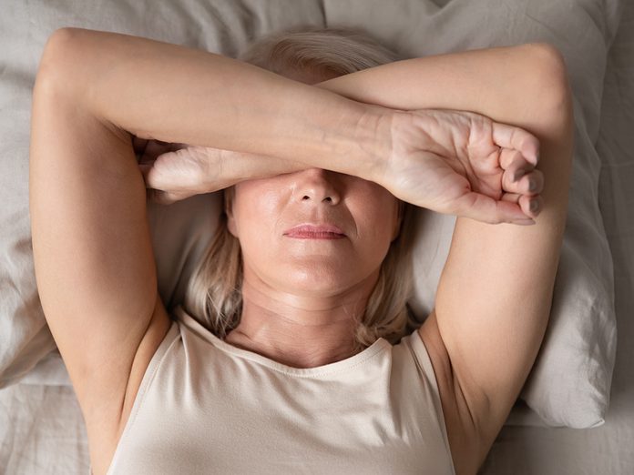 Sleepless mature woman suffering from insomnia close up, lying in bed, older female covering eyes with hands, trying to sleep, nightmares or depression, feeling headache or migraine