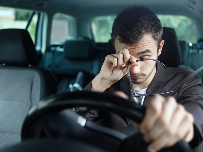 Sleepy young man rubs his eyes with his right hand. His left hand is on the steering wheel. He is sitting at his car. Road safety concept.