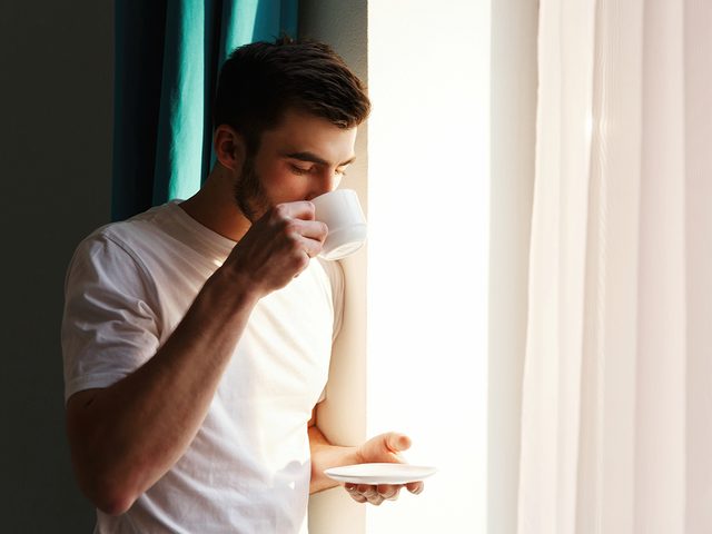 Handsome brunette bearded man stands before bright window and drinks coffee