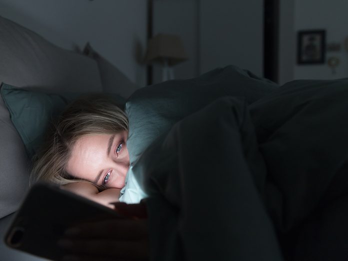 Portrait of young sleepy tired woman lying in bed under the blanket using smartphone at late night, can not sleep/ Insomnia, nomophobia, sleep disorder concept/ Dependency on a cell phone