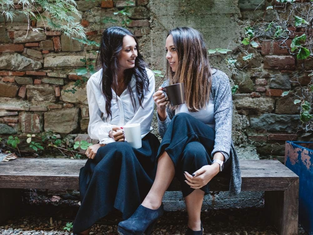 Two female friends talking in courtyard while drinking coffee