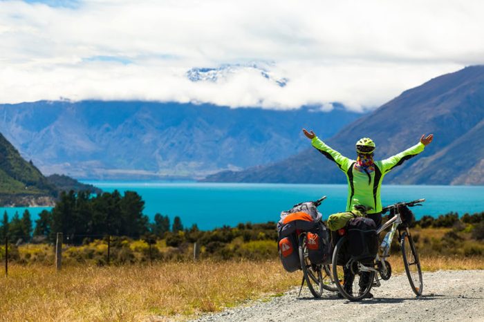 cyclist stands on the winding asphalt mountain road near Lake Pukaki view from Glentanner Park Centre near Mount Cook, on a background of blue sky with clouds, snowy Southern Alps. 