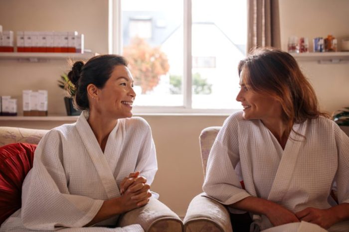 Woman friends in wellness spa waiting room, talking and smiling. Females in bathrobe sitting in health spa reception area and waiting for their spa treatment.