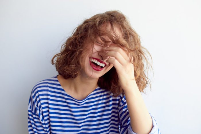 Laughing woman in marine shirt with curly hair over white wall. Toothy smile and red lips.