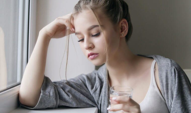 Displeased tired young beautiful girl or woman look on a pill or tablet with a glass of water near the window in white shirt and grey robe
