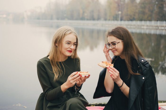 two young and stylish girls walking in the autumn park near water and eating pizza