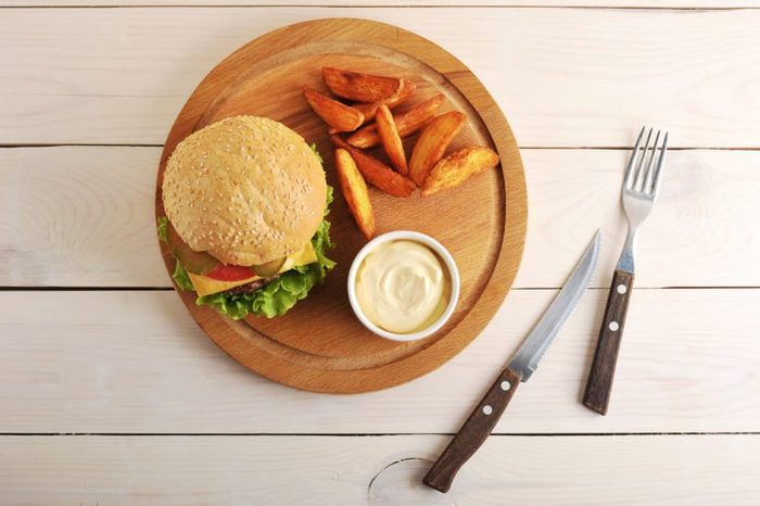 burger with Idaho potatoes and cheese sauce on a round wooden Board and a knife and fork on wooden background - top view