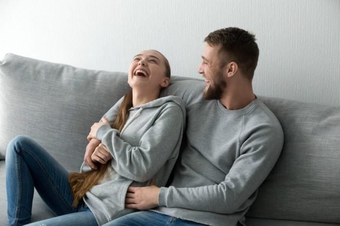 Young happy couple having fun talking laughing relaxing at home on couch, boyfriend embracing girlfriend telling funny joke sitting on sofa, humour in relationships, enjoying weekend together