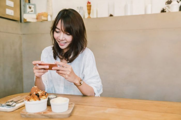 Beautiful Asian girl taking photo of chocolate toast cake, ice-cream, and milk at coffee shop. Dessert or food photograph hobby. Smartphone or mobile phone photography habit concept. With copy space
