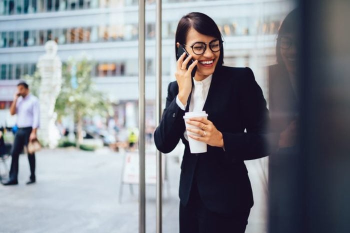 Prosperous female employee enjoying coffee break while talking on smartphone near office. Cheerful happy businesswoman in corporate wear laughing during mobile conversation outdoors in city downtown 