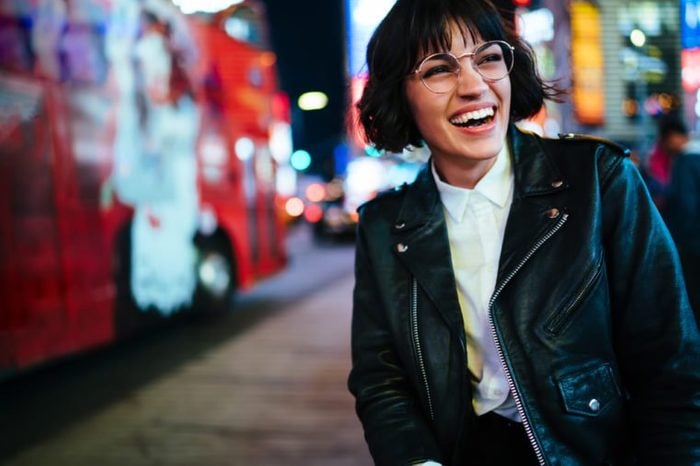 Pretty positive young woman with short haircut laughing in urban setting with night lights enjoying leisure time in New York.Happy hipster in stylish wear and eyewear having fun on megapolis street