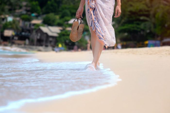 legs of woman with shawl wearing, holding in hand slippers, walking on the fine white sand of the sea beach, feeling comportable and relax in long weekend and holidays