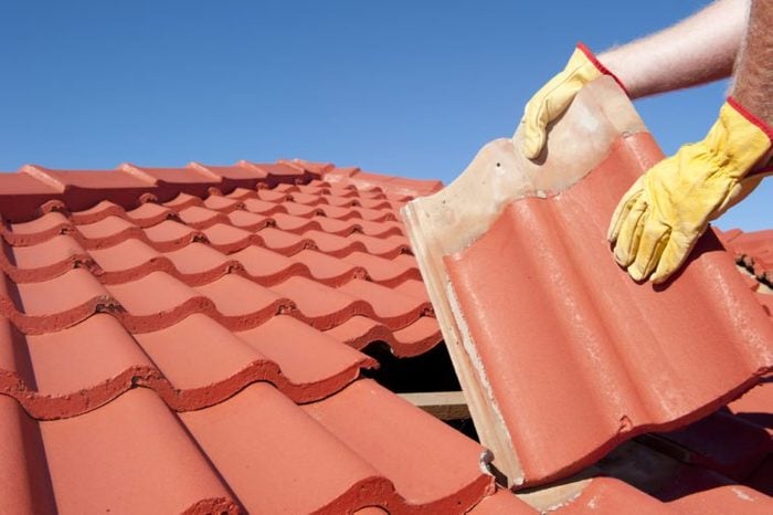 Roof repairs, worker with yellow gloves replacing red tiles or shingles on house with blue sk
