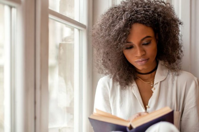 Beautiful african american young woman sitting by the window and reading a book.