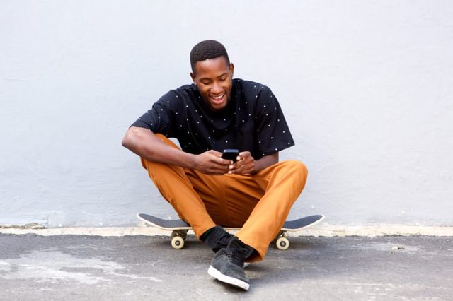 Portrait of attractive young african man sitting with skateboard and using smart phone