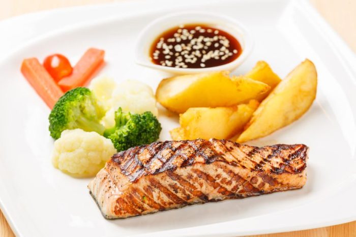 Salmon grilled steak served with fried potato carrot, broccoli, cauliflower, sweet sauce and white sesame in ceramic dish on wooden table