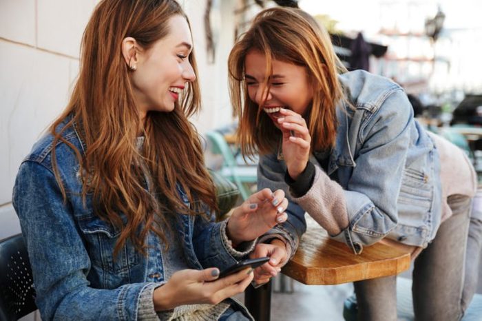 Two young pretty girls in casual stylish wear laughing while sitting at cafe