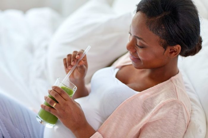 pregnancy, people and rest concept - close up of happy pregnant african american woman drinking green vegetable juice or smoothie in bed at home