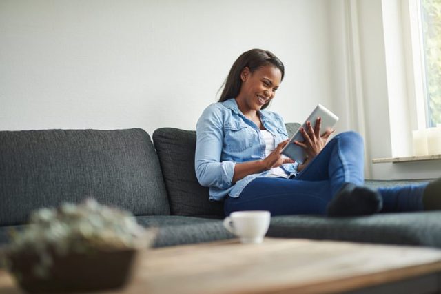 Young African woman laughing while relaxing on her living room sofa browsing online with a digital tablet