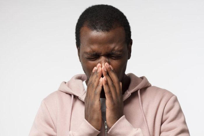 Body language. Sick young african man in pink hoodie covering face with hands; sneezing; isolated on gray background