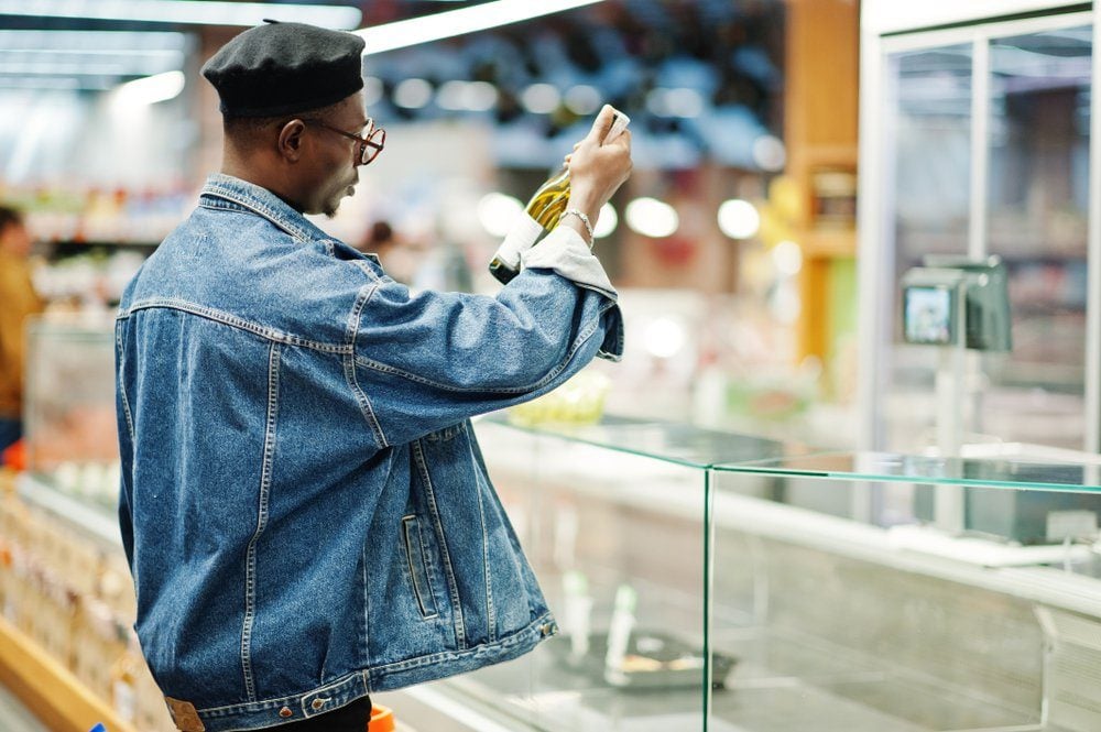 Stylish casual african american man at jeans jacket and black beret holding basket and looking on bottle of wine, shopping at supermarket.