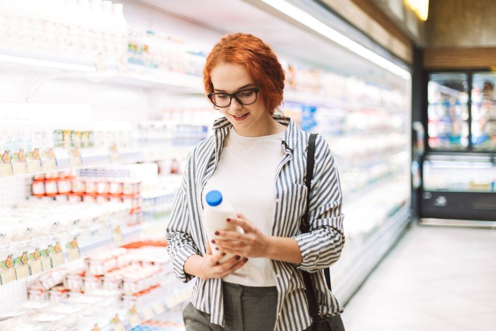 Pretty smiling girl in eyeglasses and striped shirt dreamily looking on bottle of milk in hands in modern supermarket