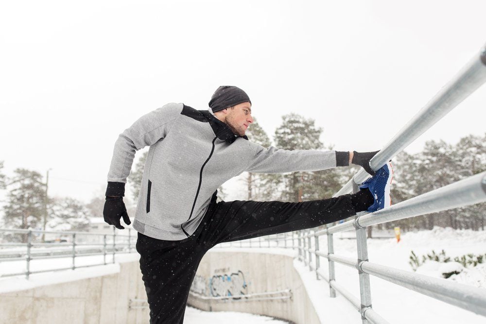 fitness, sport, people, exercising and healthy lifestyle concept - young man stretching leg and warming up at fence in winter