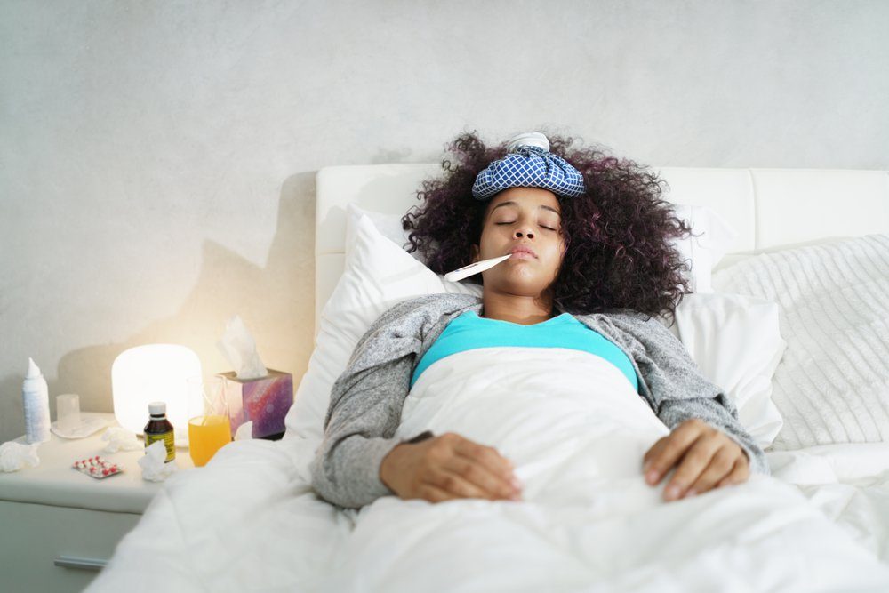 Sick african american girl with flu in bedroom at home. Ill young black woman with cold, lying in bed and holding a thermometer in her mouth.
