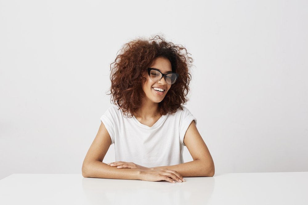 Beautiful african girl in glasses laughing sitting over white background. Copy space.