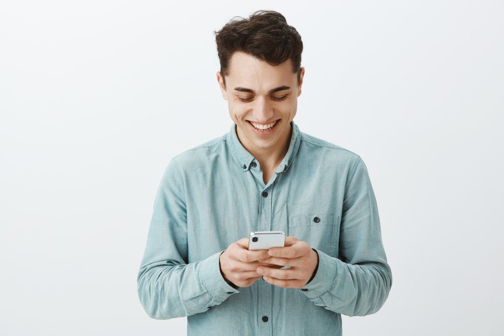 Portrait of pleased carefree handsome guy with short dark hair in shirt, holding smartphone and laughing with broad smile, texting friend funny meme or relaxing watching video in internet