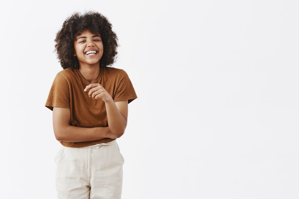 Studio shot of cute african american woman with afro hairstyle in trendy summer outfit having fun laughing out loud from joy and funny joke gesturing with palm chuckling and smiling carefree at camera