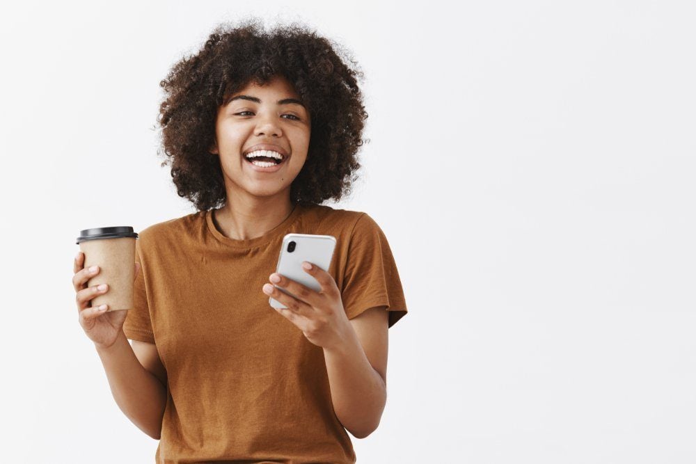 Waist-up shot of trendy carefree african american girl with curly hair in brown t-shirt laughing while talking with friends drinking coffee from paper cup and holding smartphone over gray background