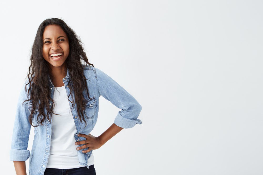 People, leisure and lifestyle concept. Good-looking young Afro-American woman with long wavy hair wearing stylish clothing smiling broadly, laughing happily at someone`s joke, having fun indoors.
