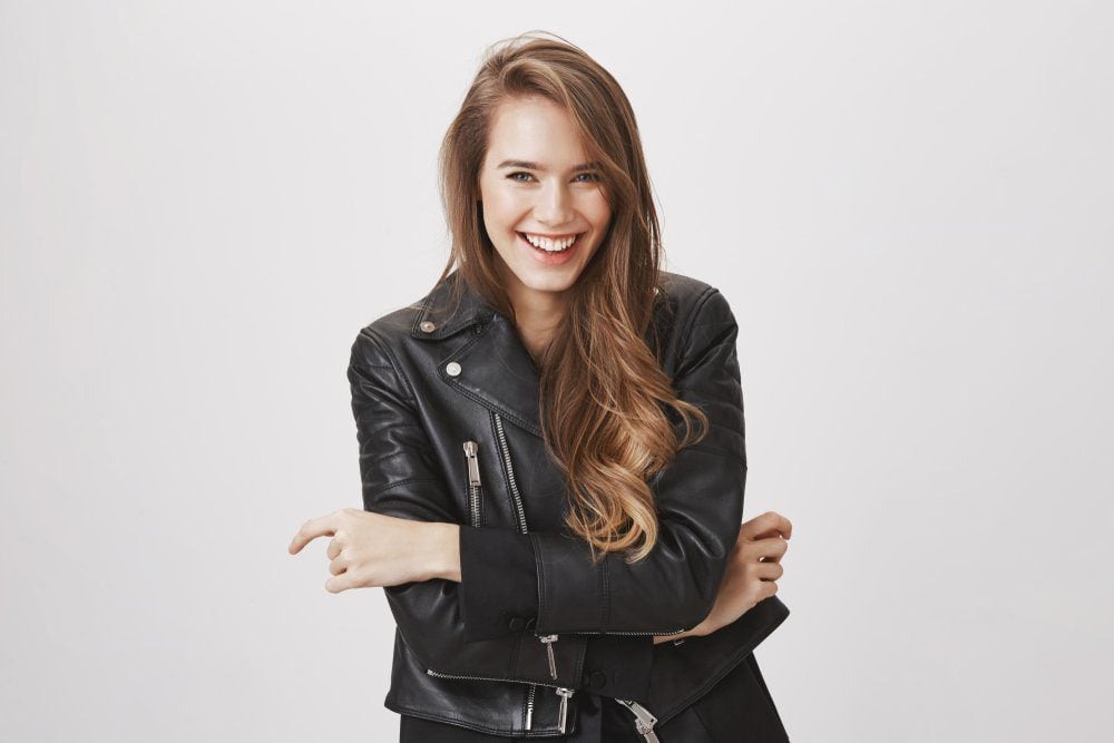 Laughing out loud in this beautiful spring day. Portrait of attractive urban girl with blond hair in leather jacket holding hands on chest, chuckling, looking at camera with happy playful expression