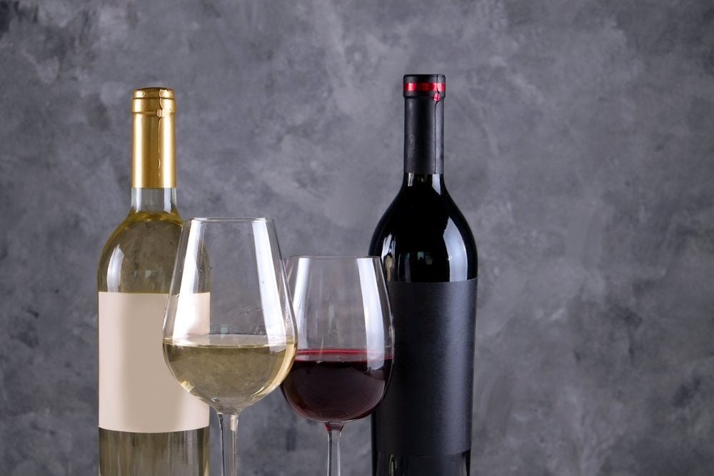 Two bottles of red and white wine with matte black & white blank labels on concrete wall background. Wineglasses half full with vintage expensive wine of different type. Close up, copy space.
