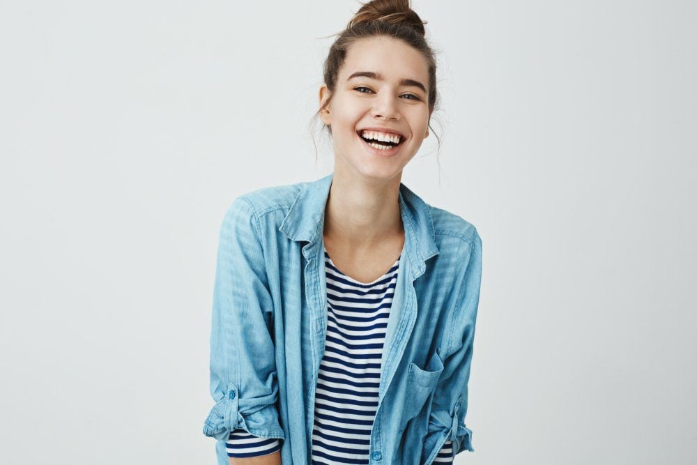 Girl likes funny jokes. Smart good-looking student with bun hairstyle trembling from laugh, smiling positively and being in great mood while standing over gray background. Woman watch hilarious show