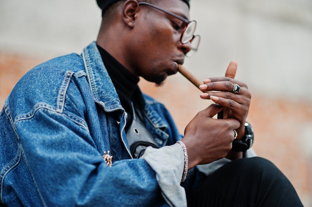 African american man in jeans jacket, beret and eyeglasses, lights a cigar.