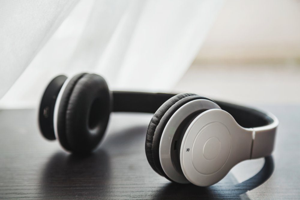 White minimalist wireless headset placed on a dark wooden laminated surface with blurred white curtain crossing a window behind it