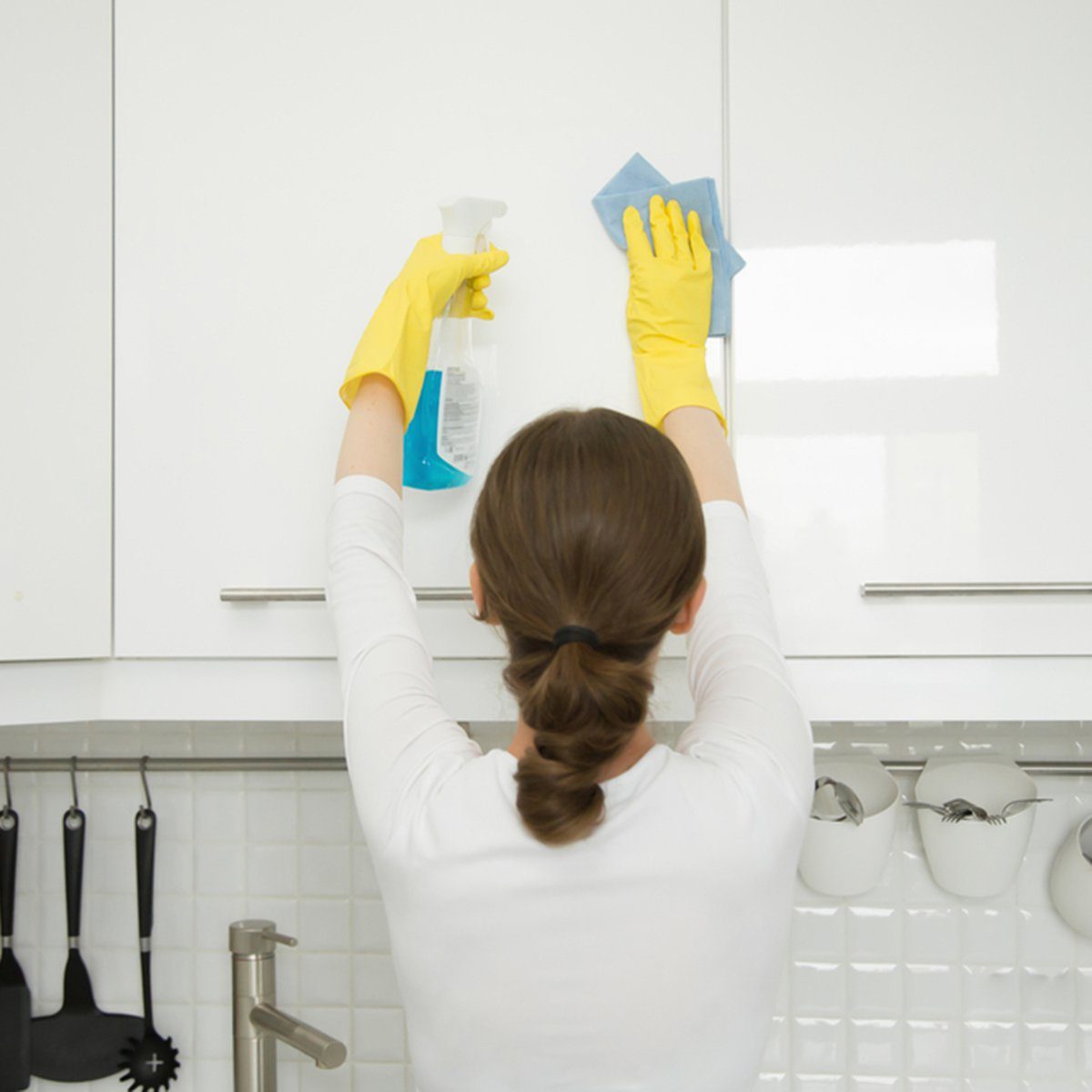 Rear view at an attractive young woman cleaning a surface of white kitchen wall cabinet, wearing rubber protective yellow gloves, with rag and spray bottle detergent.