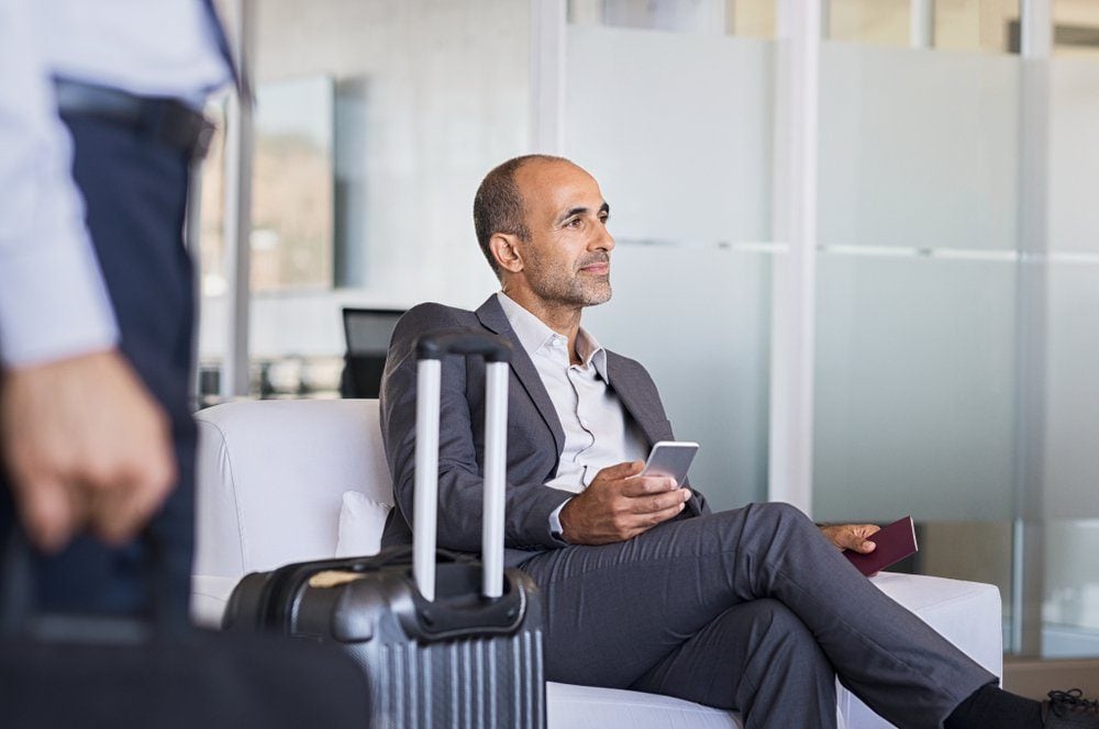Mature businessman expecting airplane at the airport. Thoughtful business man waiting for flight in airport. Formal business man sitting in airport waiting room with luggage and phone in hand.