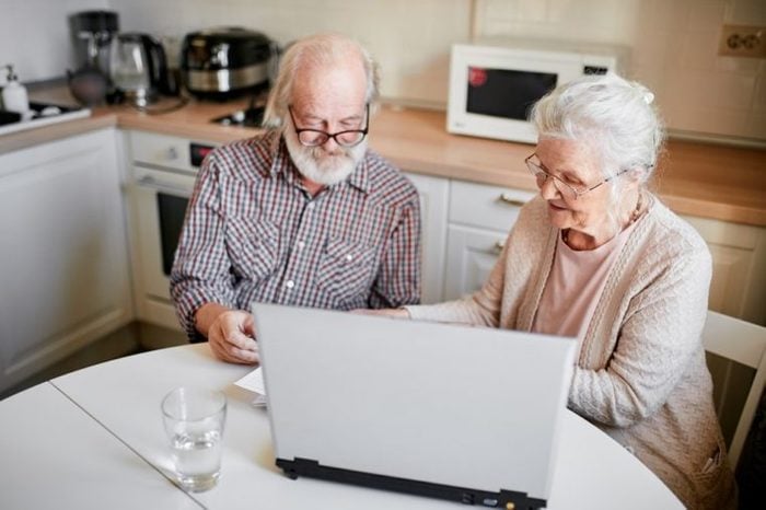 Senior couple at home kitchen using laptop, writing email to their foreign far-away friends, one of the pensioners holding document, another typing on computer keyboard. Close up.