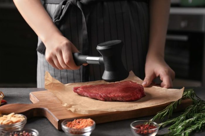 Woman beating raw steak with meat mallet in kitchen
