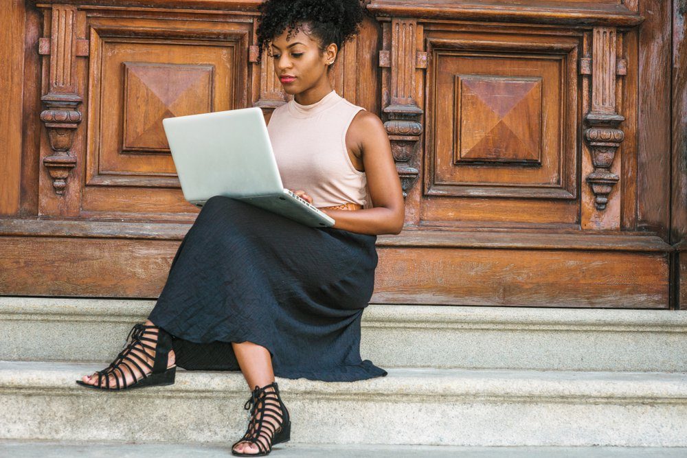 African American Student studying in New York, with afro hairstyle, wearing sleeveless light color top, black skit, strappy sandals, sitting by office door in New York, working on laptop computer.