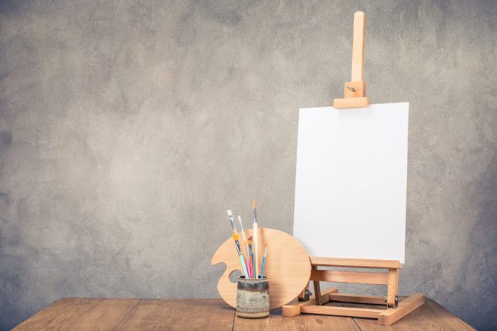 Portable desk easel for painting with canvas blank, brushes and artist's palette on wooden table front concrete wall background. Retro style filtered photo