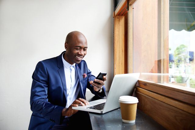Portrait of happy african businessman using phone while working on laptop in a restaurant