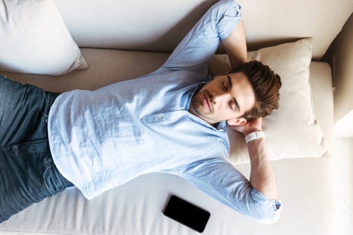 Top view of a young man sleeping on the couch at home with mobile phone