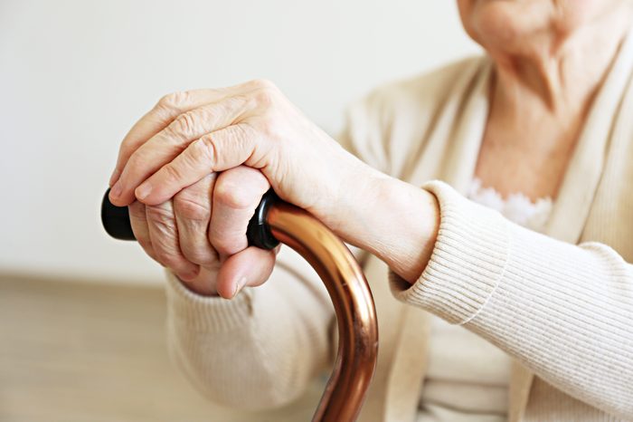 Elderly woman sitting in nursing home room holding walking quad cane with wrinked hand. Old age senior lady wearing beige cardigan, metal aid stick handle bar close up. Interior background, copy space