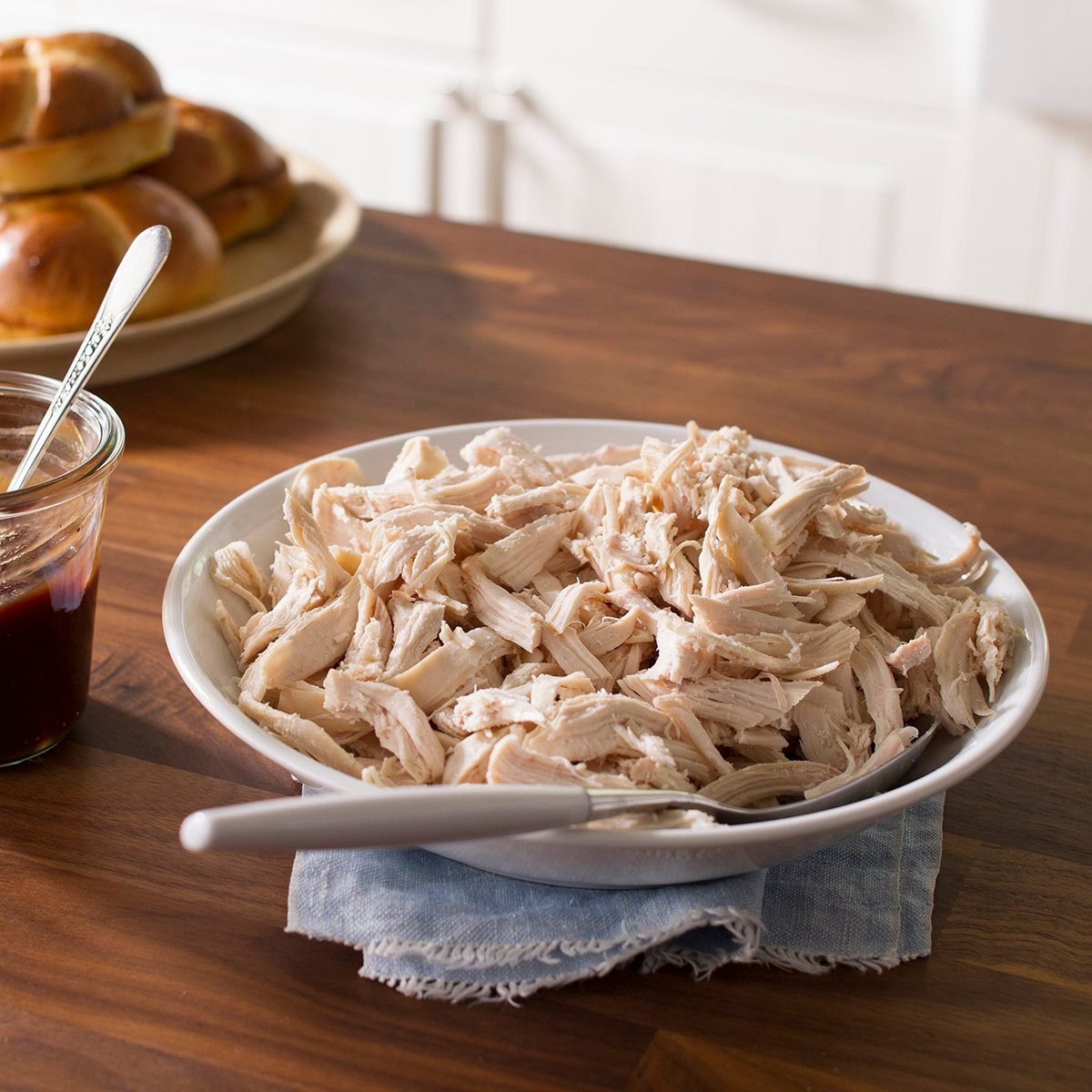 A white plate filled with shredded chicken on a wooden table beside a plate of buns and jar of gravy