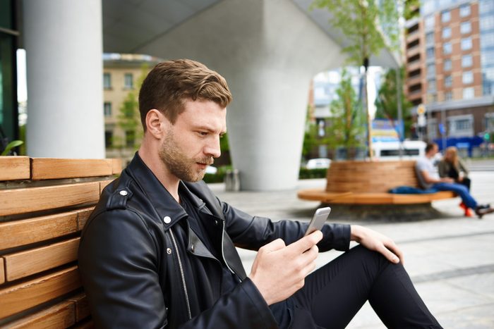 rude conversation habits - Caucasian attractive man use mobile for internet access when waiting for a girl Male checking email box in the city on bench. modern guy in leather jacket watch video online or write in social network