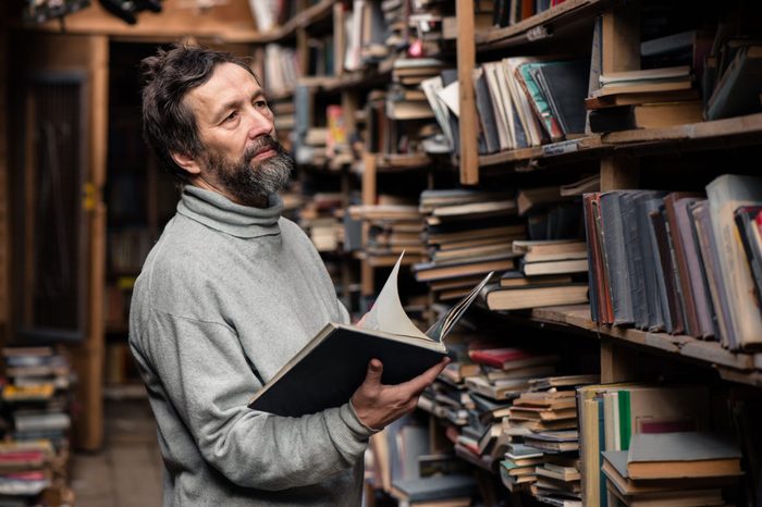 Portrait of authentic senior man with beard and good eyes reading book in the hand on bookshelf background